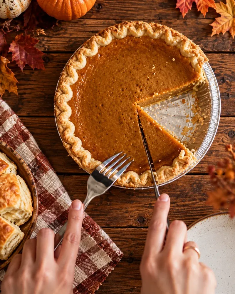 Fork cutting into a pumpkin pie on a wooden table with leaves and a glass of cranberry juice