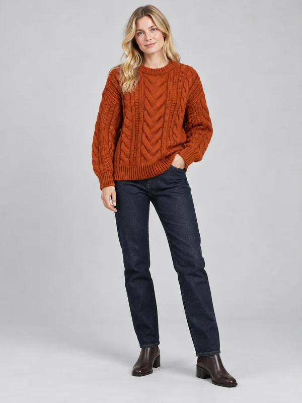 AI model in a rust cable-knit sweater and dark jeans on a gray studio floor