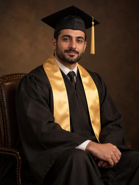 Seated graduate in a black gown and gold stole in a moody studio